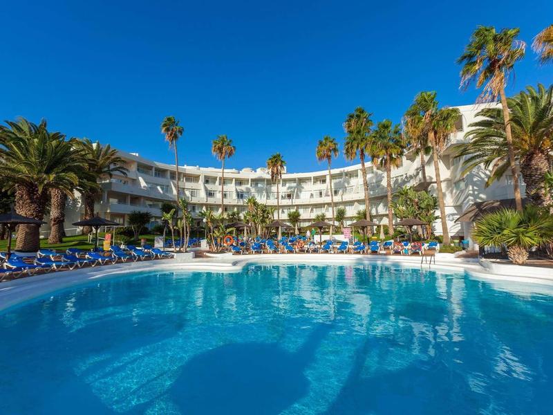 Piscina azzurra davanti a un hotel con palme sotto un cielo turchese.