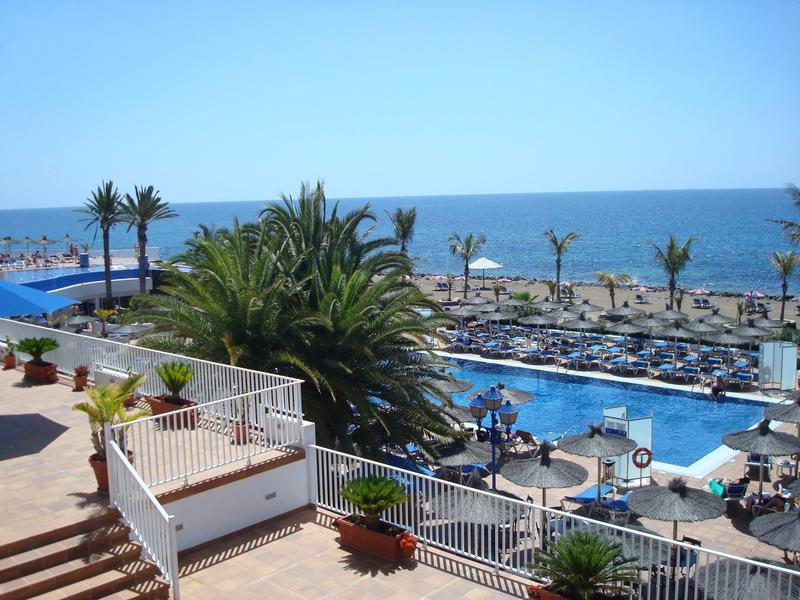 Piscina dell'hotel con lettini, palme e vista sul mare con cielo limpido