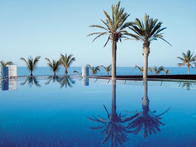 Camera d'hotel luminosa con letto matrimoniale, balcone e vista su palme e mare.