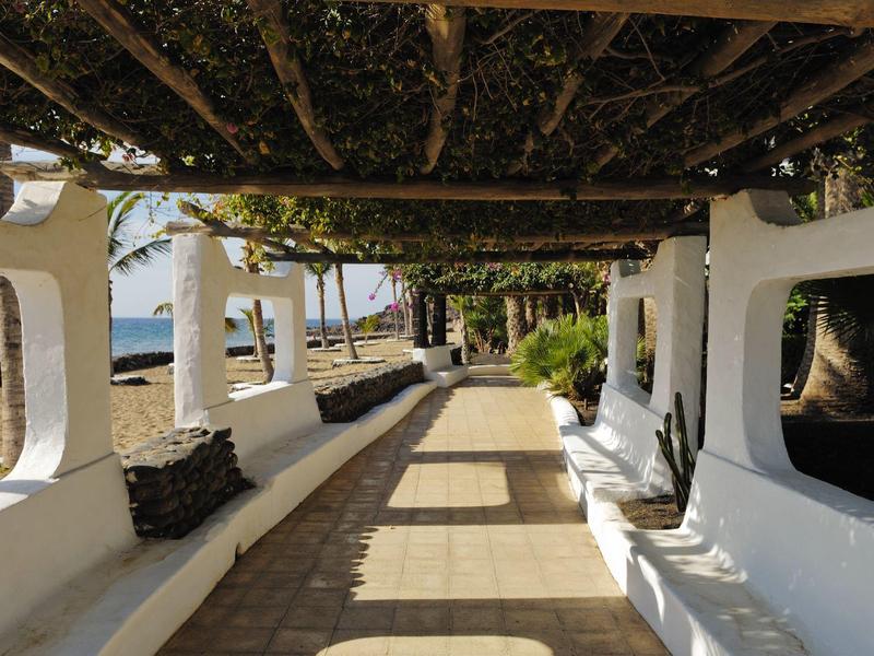 Passaggio esterno coperto con panchine bianche e cuscini, circondato da alberi con vista mare.