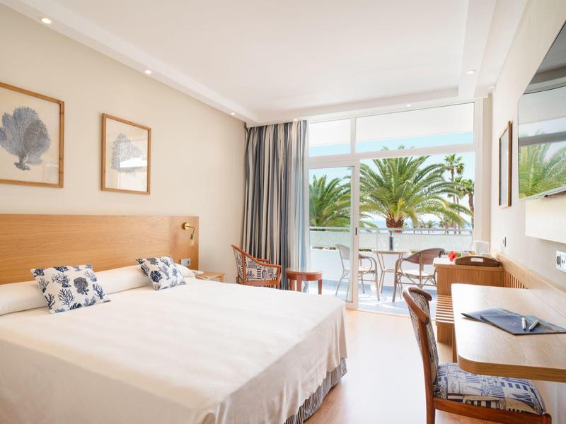 Camera d'hotel luminosa con letto matrimoniale, balcone e vista su palme e mare.