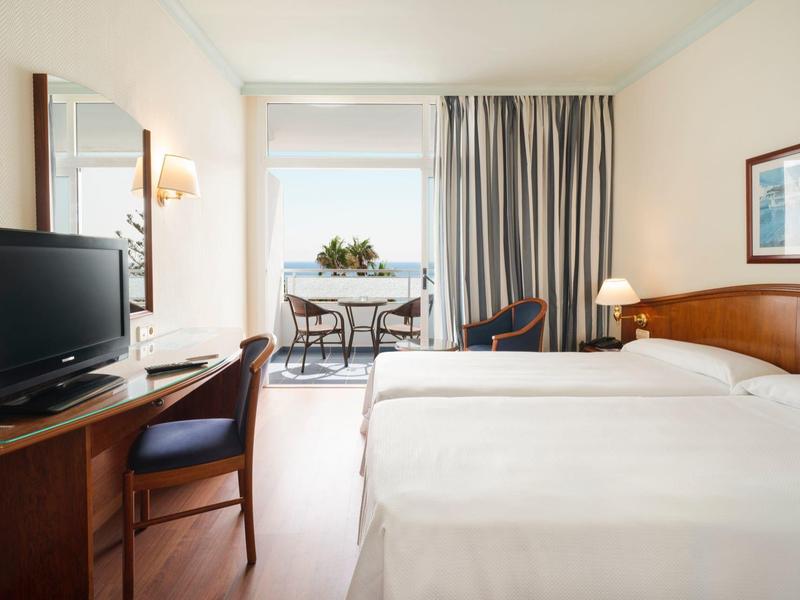 Camera d'albergo luminosa con due letti, una scrivania, una TV e un balcone.