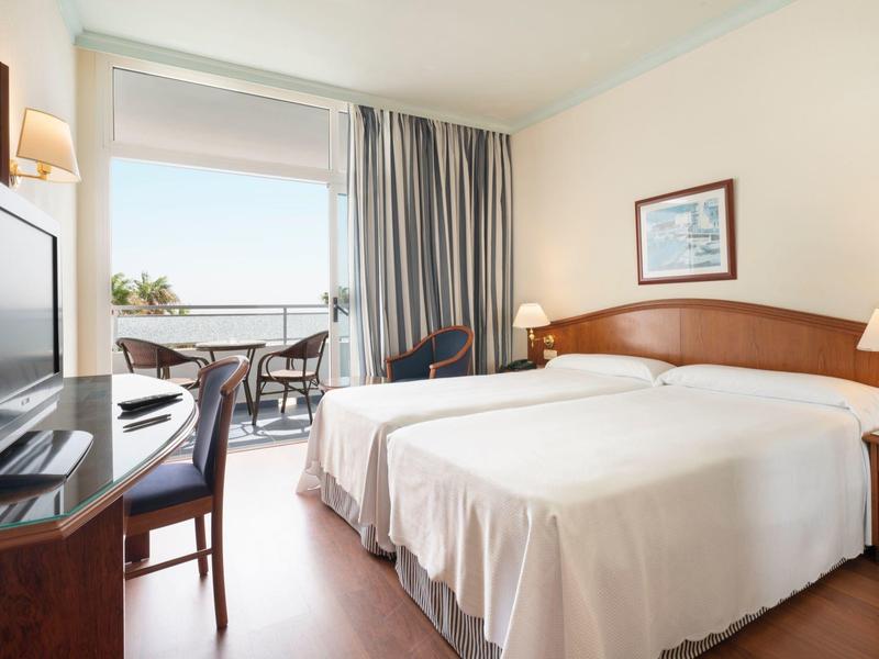 Camera d'albergo luminosa con due letti, TV, scrivania e balcone con vista mare.