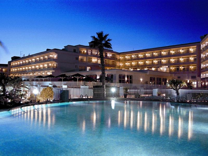 Edificio dell'hotel illuminato con grande piscina esterna di notte e palme.