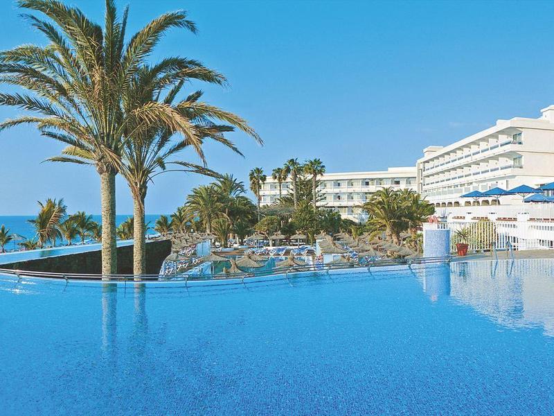 Grande piscina a sfioro con palme vicino a un hotel di lusso sul mare con cielo sereno.