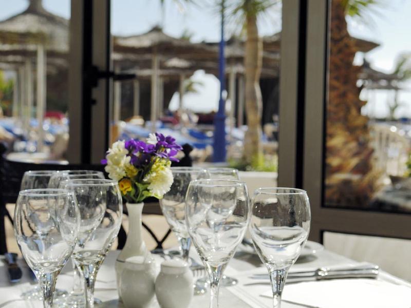 Tavolo elegantemente apparecchiato con bicchieri e fiori davanti a una finestra con vista su palme e spiaggia.