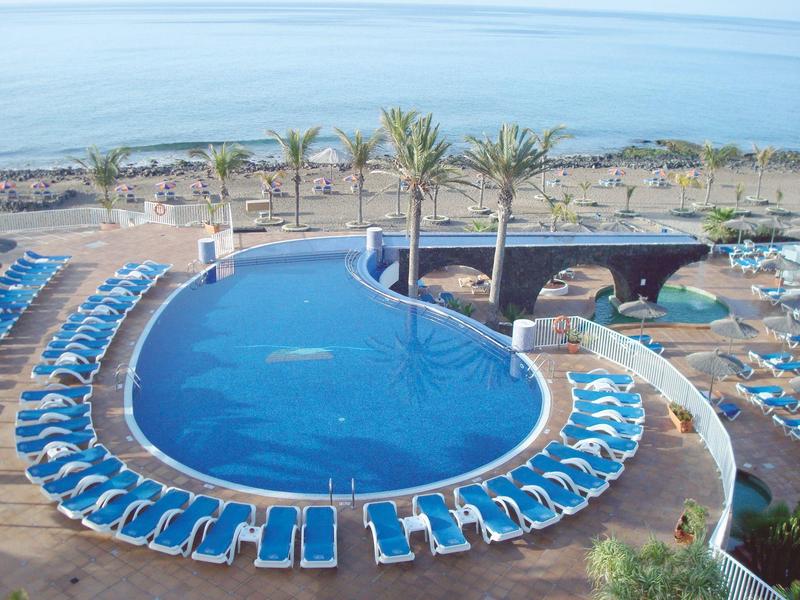 Piscina rotonda dell'hotel con lettini blu, palme e vista mare sullo sfondo.