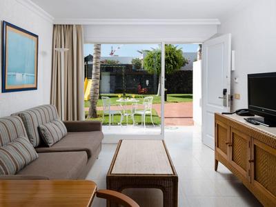 Stanza luminosa con divano, mobili in legno, TV e porta che si apre su giardino con sedie e tavolo.