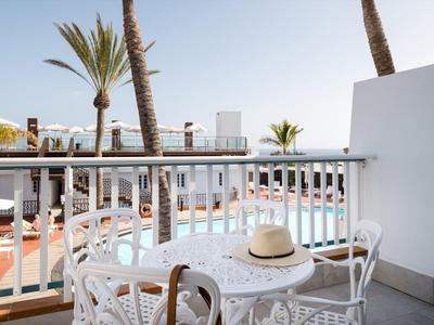 Balcone con tavolo e sedie bianche che si affaccia su piscina e palme sotto cielo sereno