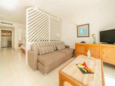 Soggiorno luminoso con divano beige, mobili in legno e TV, separato da una parete bianca.