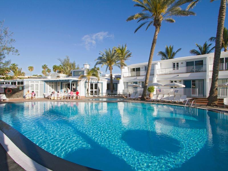 Piscina esterna luminosa dell'hotel con palme e edifici bianchi sotto un cielo azzurro.