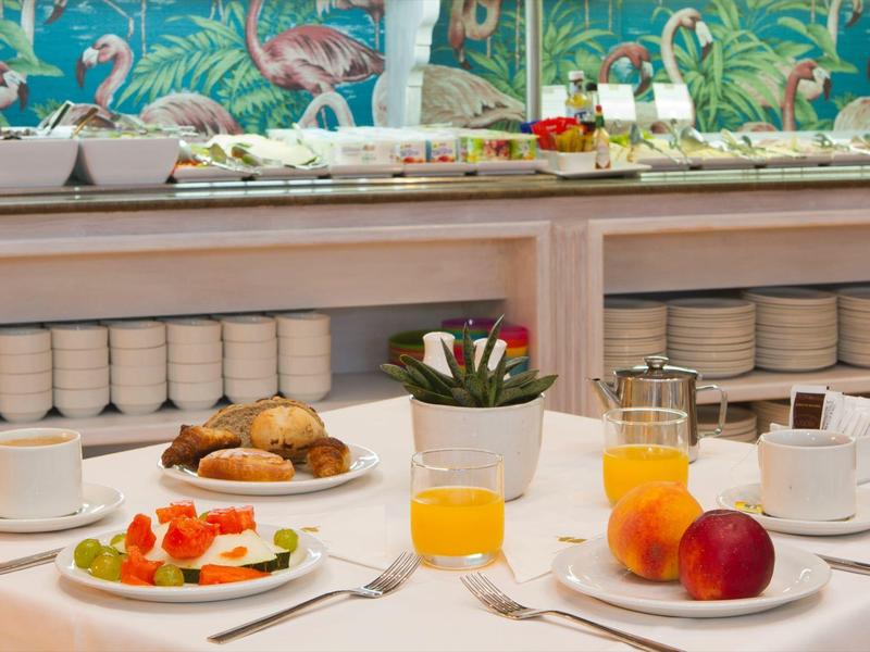 Tavolo apparecchiato con colazione, dolci, frutta, succo e caffè davanti a carta da parati tropicale.