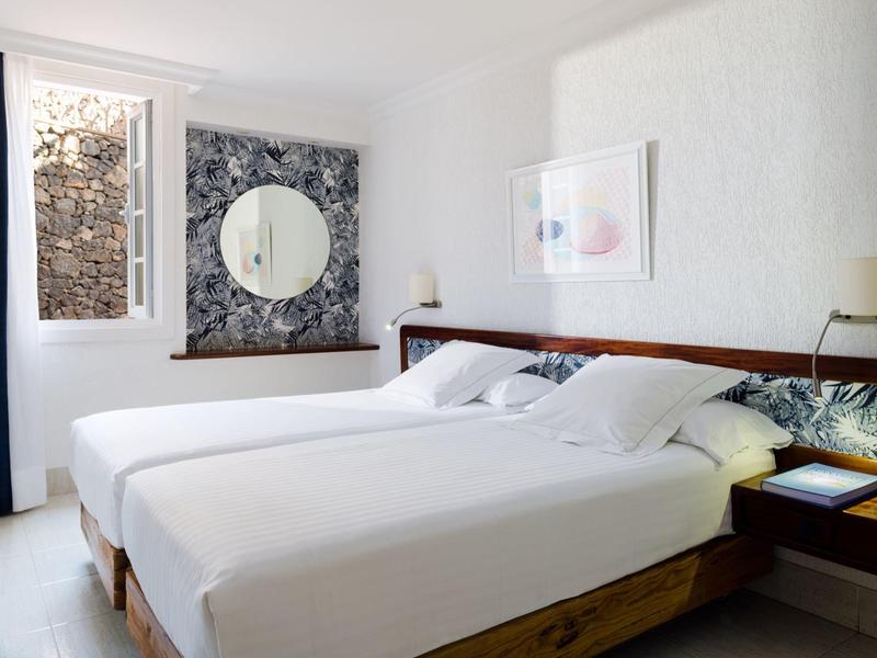 Camera d'albergo luminosa con letti singoli, arredamento moderno e grande specchio su carta da parati floreale.