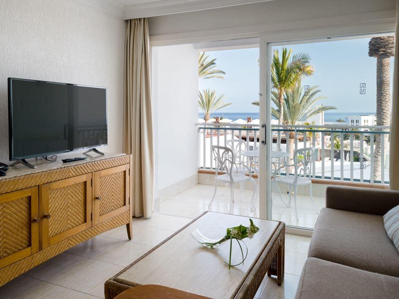 Soggiorno luminoso con divano, tavolo in legno, TV su mobile in legno e balcone con vista palme.