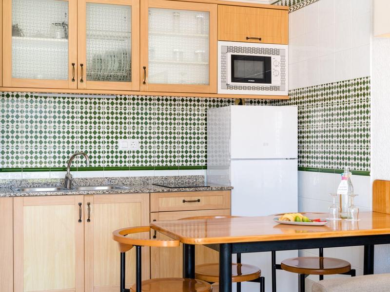 Piccola cucina con mobili in legno, frigorifero bianco, microonde e tavolo in legno con sgabelli.