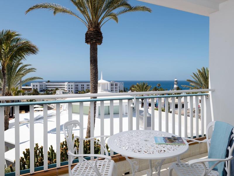 Balcone con posti a sedere che si affaccia su palme, edifici bianchi e oceano blu sotto un cielo limpido.