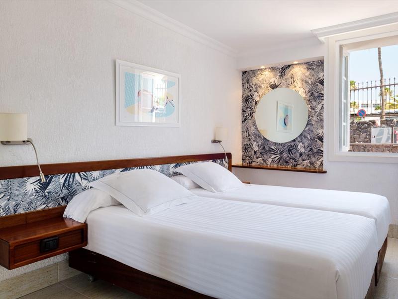 Camera con due letti singoli, biancheria bianca, testiera in legno e una finestra che fa entrare la luce.