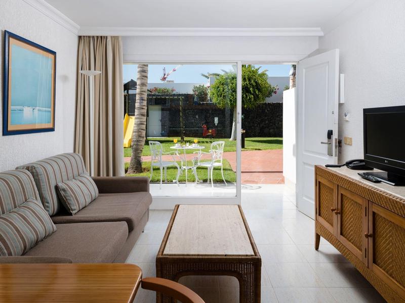 Stanza luminosa con divano, mobili in legno, TV e porta che si apre su giardino con sedie e tavolo.