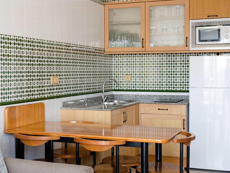 Cucina compatta con mobili in legno, piastrelle verdi, piccolo tavolo e frigorifero bianco.