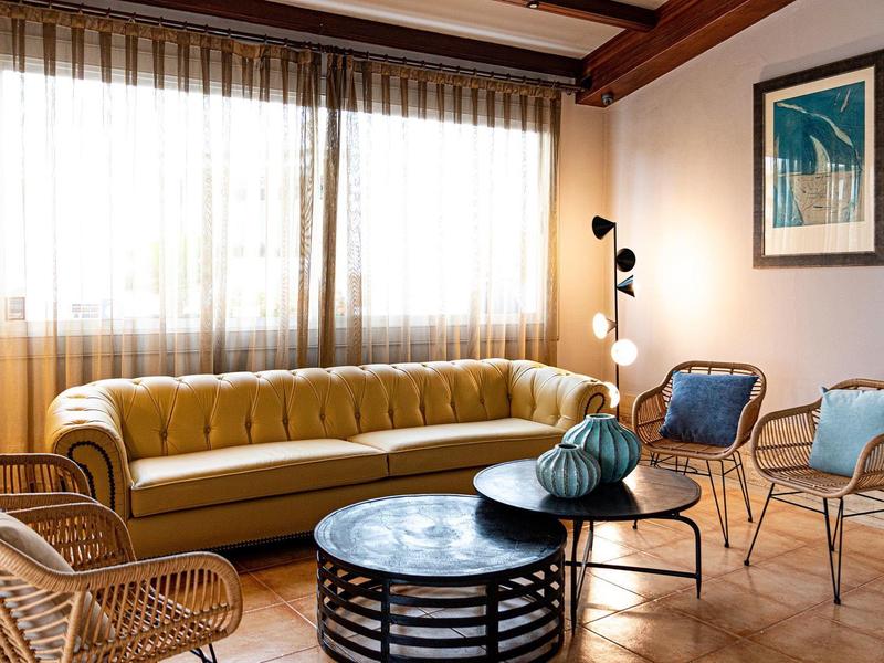 Accogliente lounge dell'hotel con divano color senape, sedie in rattan, tavolino rotondo nero e grandi finestre.