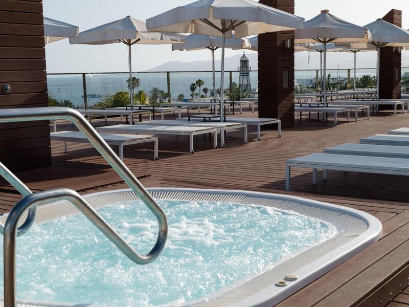 Vasca idromassaggio esterna con lettini bianchi e ombrelloni su terrazza in legno con vista sul mare.