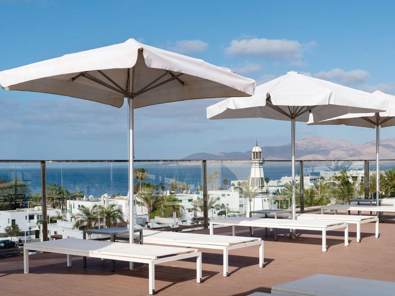 Lettini bianchi con ombrelloni grandi vicino a una piscina con vista sul mare e sugli edifici.
