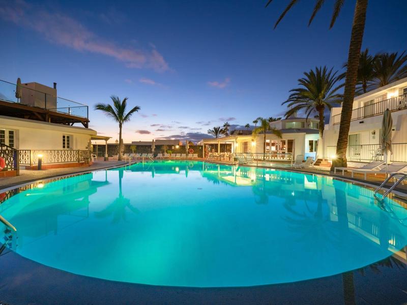 Piscina rotonda illuminata al tramonto circondata da edifici bianchi e palme.