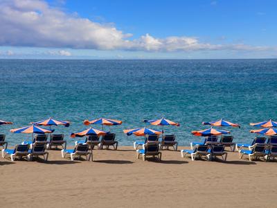 Strand mit mehreren blauen Sonnenschirmen und Liegestühlen vor ruhigem, blauem Meer unter blauem Himmel.