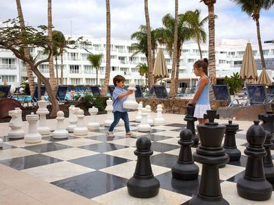 Zwei Kinder spielen riesiges Schachspiel im Freien vor weißem Hotel und Palmen.