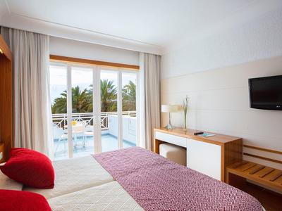 Helles Hotelzimmer mit Doppelbett, roten Kissen, Balkon und Blick auf Palmen und Pool.
