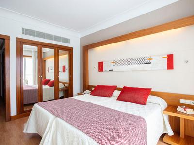 Modernes Schlafzimmer mit großem Bett, roten Kissen, Holzmöbeln und Spiegeln in heller Atmosphäre.