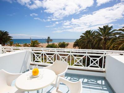 Balkon mit weißem Geländer, Tisch, zwei Stühlen, Saft, Palmen und Meer unter blauem Himmel.