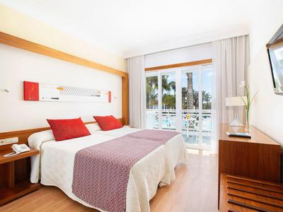Hell eingerichtetes Hotelzimmer mit Doppelbett, roten Akzenten und Balkon mit Blick auf Palmen.