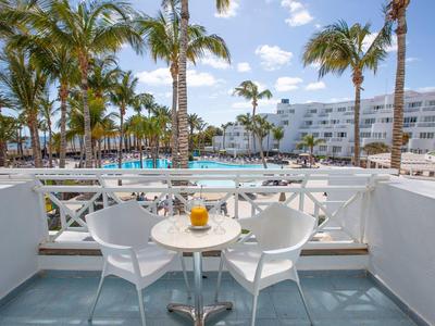 Balkon mit zwei weißen Stühlen und kleinem Tisch, Palmen und Hotelgebäude im Hintergrund.
