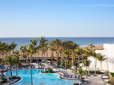 Hotelanlage mit großem blauem Pool, Palmen und Blick auf das ruhige blaue Meer unter blauem Himmel.