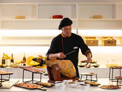 Ein Koch schneidet Fleisch von einem großen Schinken in einer modernen Küche mit weißen Regalen.