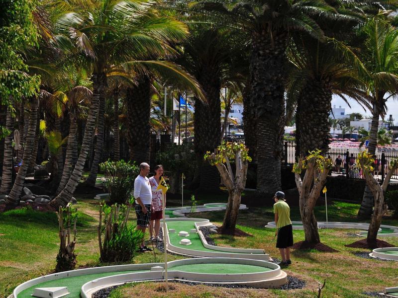 Persone che giocano a mini golf in un percorso fiancheggiato da palme in una giornata di sole.