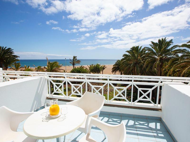 Balcone con mobili bianchi che si affaccia su palme e mare sotto un cielo azzurro.
