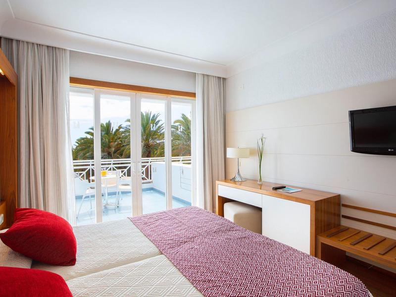 Camera d'hotel luminosa con letto matrimoniale, cuscini rossi, balcone e vista su palme e piscina.