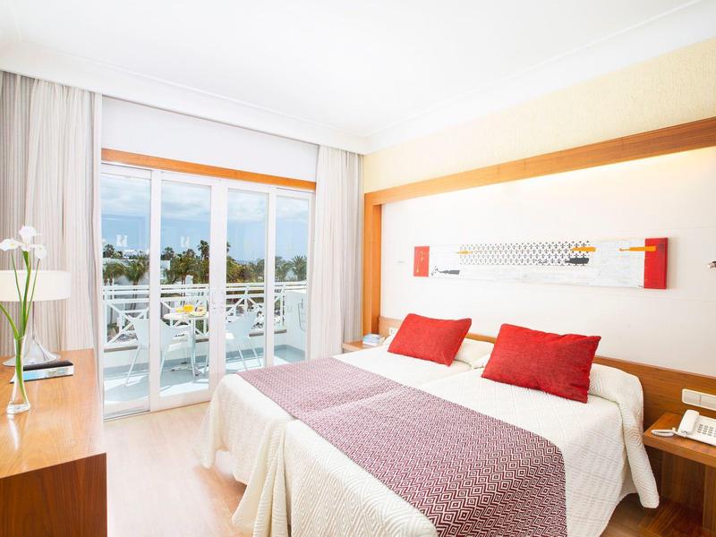 Camera d'hotel luminosa con letto matrimoniale, accenti rossi e balcone con vista sull'acqua.