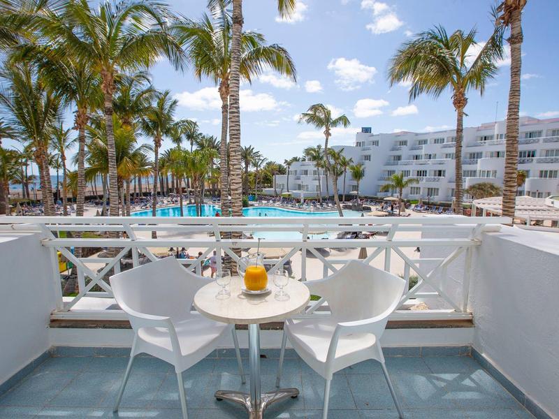 Balcone con tavolo per due, vista sulla piscina, palme e edificio alberghiero bianco sotto un cielo blu.