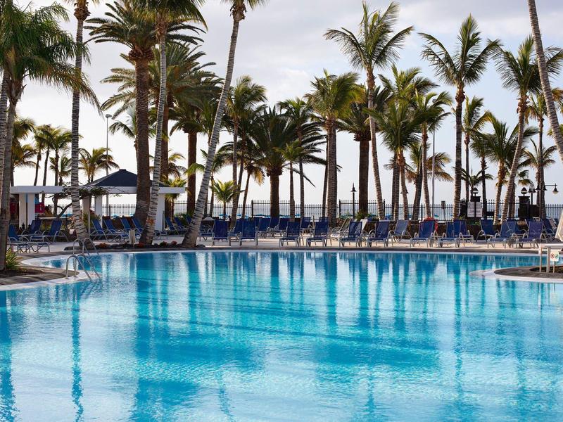 Grande piscina con acqua azzurra limpida, circondata da ombrelloni e palme sotto un cielo nuvoloso.