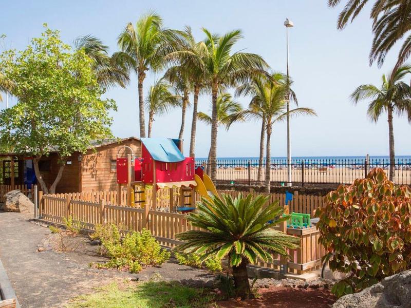 Area giochi per bambini con casette, palme, cielo sereno e vista sulla spiaggia.