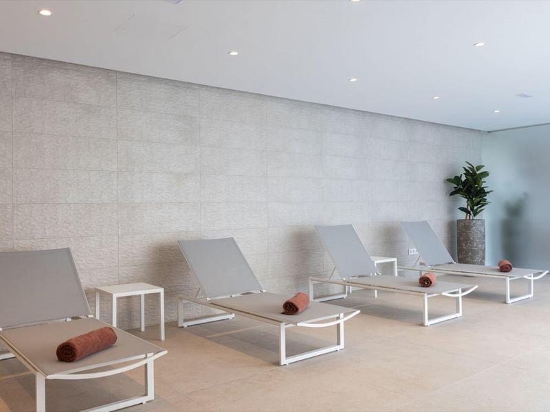 Area relax moderna con lettini grigi e tavolini bianchi davanti a una parete chiara.