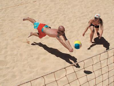 Dwóch zawodników gra w siatkówkę plażową, jeden skacze po piłkę.
