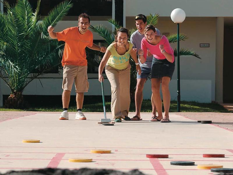 Groupe de quatre personnes jouant au shuffleboard devant un hôtel avec des palmiers.