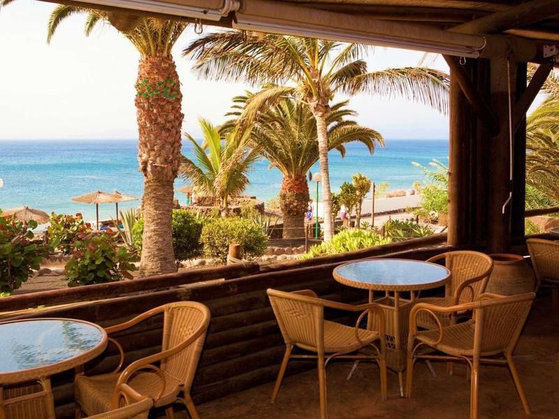 Terrasse avec tables et chaises donnant sur des palmiers et la mer en arrière-plan.