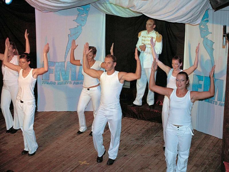 Sept personnes exécutent une danse sur scène portant des costumes clairs sur un sol en bois.