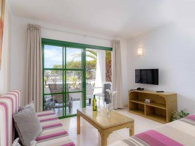 Helles Zimmer mit Sofa, Holz-Couchtisch, TV und Balkon mit Blick auf Palmen und Meer.