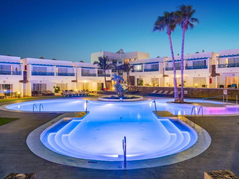 Moderner Hotelpool mit Beleuchtung, zwei Palmen, niedrigen Gebäuden und klarem Abendhimmel.